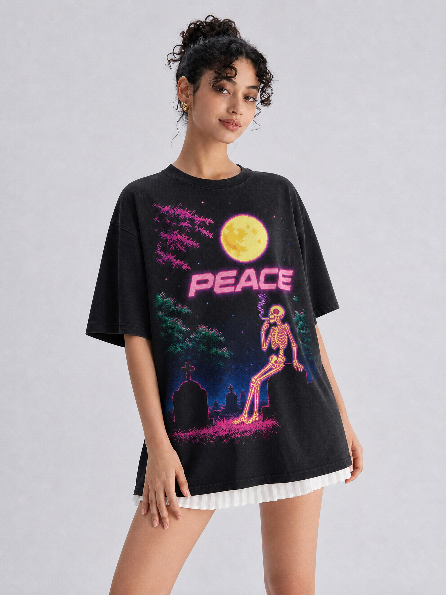 A woman modeling an oversized black graphic T-shirt with a colorful retro design featuring a yellow moon, pink 'PEACE' text, a skeleton, and a graveyard scene, paired with a white pleated skirt against a light gray studio background.