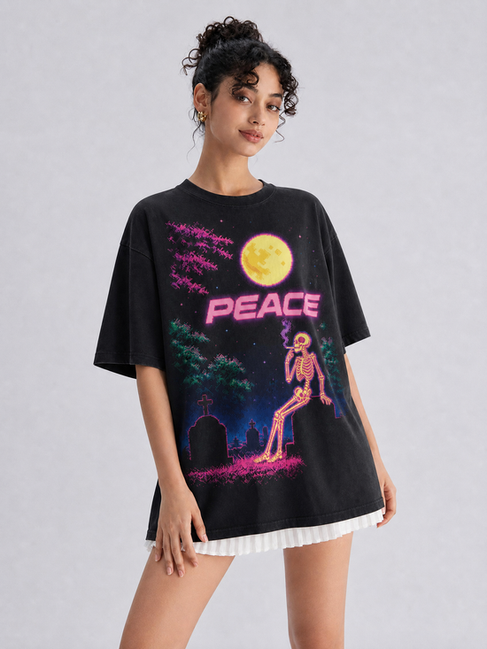 A woman modeling an oversized black graphic T-shirt with a colorful retro design featuring a yellow moon, pink 'PEACE' text, a skeleton, and a graveyard scene, paired with a white pleated skirt against a light gray studio background.