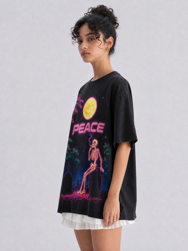 A model wearing an oversized black graphic t-shirt with a neon-style print of a skeleton, a yellow moon, and the word 'PEACE' in pink, paired with a white ruffled skirt, against a light gray studio background.
