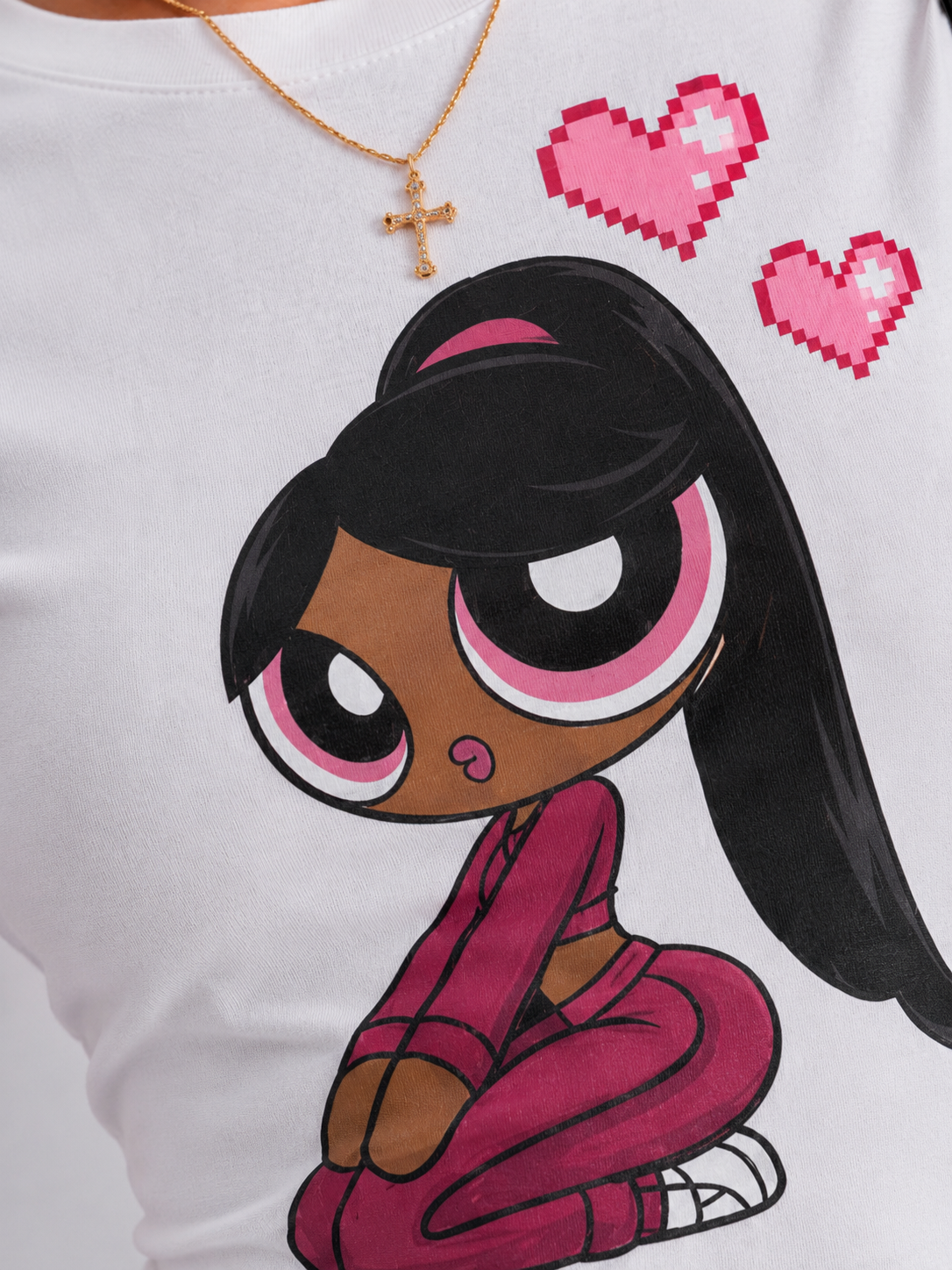A close-up of a white t-shirt featuring a cartoon graphic of a girl with long black hair, brown skin, and a pink outfit, with two pink pixel hearts above her. A person wearing a gold cross necklace is partially visible.