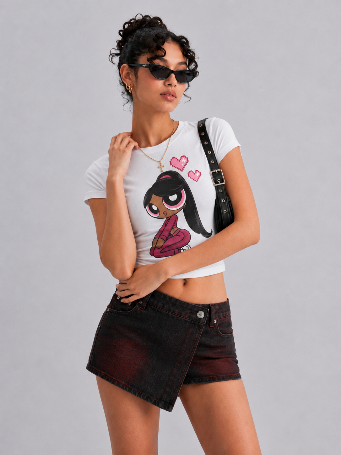 A model wearing a white short-sleeve crop top with a graphic of a cartoon girl and pink hearts, paired with a dark denim asymmetrical mini skirt and a black shoulder bag, against a solid gray background.