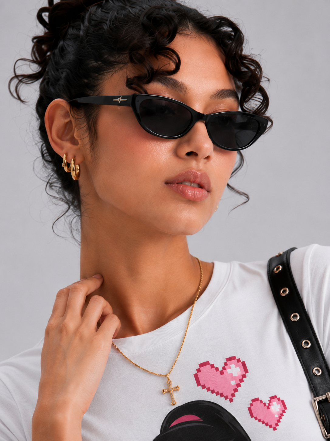 A close-up of a model wearing black cat-eye sunglasses with a small gold logo on the temple, styled with a white graphic t-shirt, gold jewelry, and a black shoulder strap against a solid gray background.