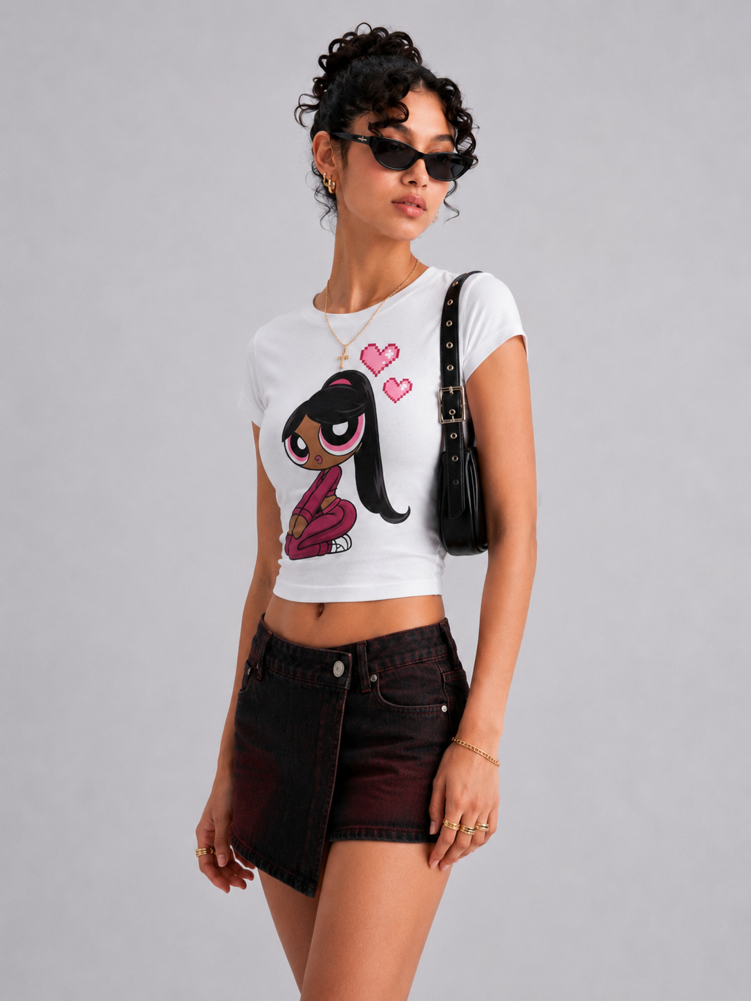 A model wearing a white short-sleeve crop top with a cartoon graphic of a girl with pink hair and hearts, paired with a dark denim mini skirt and a black shoulder bag, against a solid gray background.