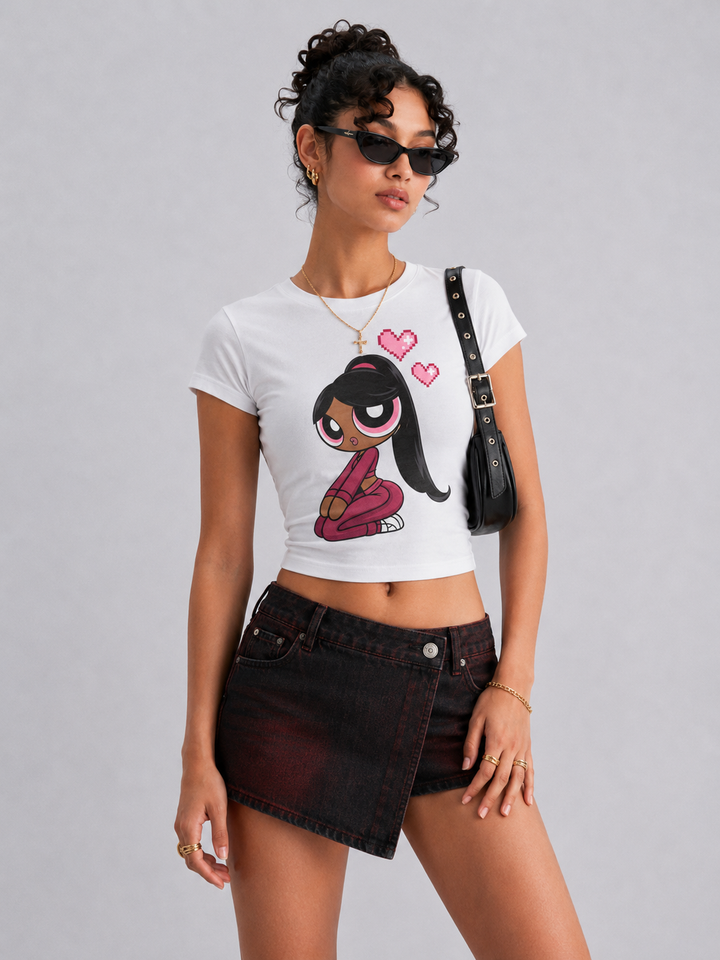 A model wearing a white short-sleeve crop top t-shirt with a cartoon graphic of a girl with pink hair and hearts, paired with a dark denim asymmetrical mini skirt and a black shoulder bag, against a solid gray background.