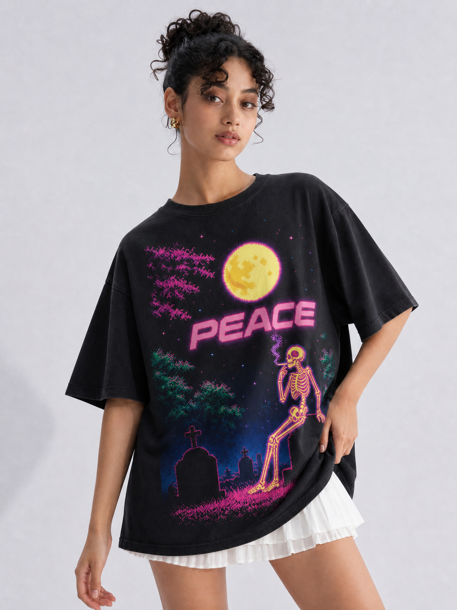 A woman modeling an oversized black graphic t-shirt with a colorful neon-style print featuring a skeleton, a moon, trees, and the word 'PEACE' in pink. She is wearing a white pleated skirt against a light gray studio background.