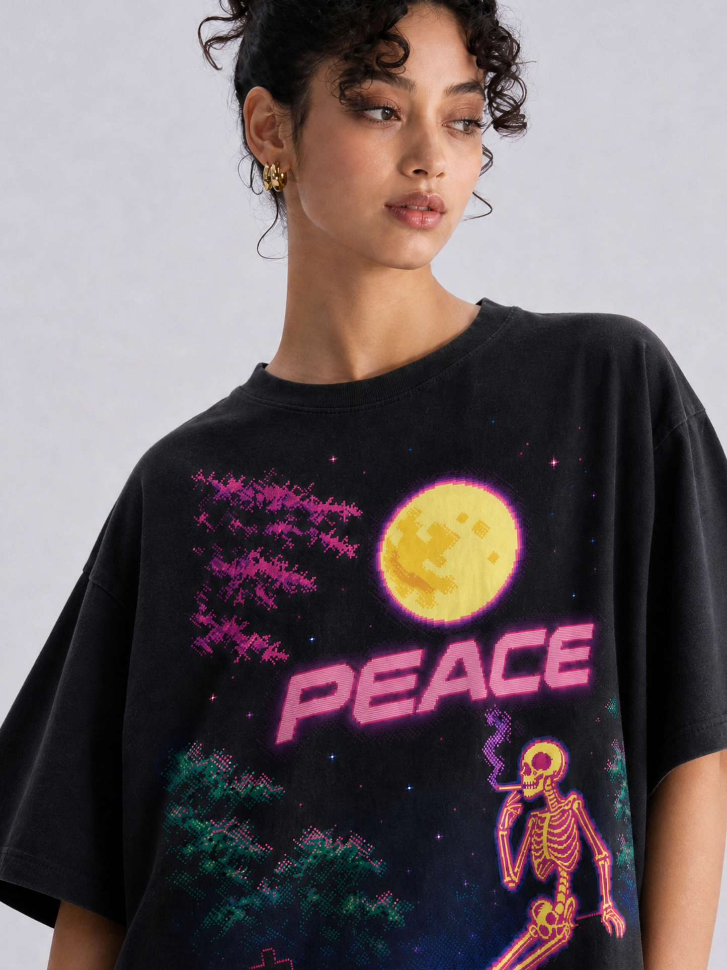 A woman wearing a black oversized graphic t-shirt with a colorful neon-style print featuring a skeleton smoking, a full moon, and the word 'PEACE'.